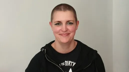 A photo of the author Lynda Clark standing in front of a white background. She is wearing black and has a shaved head.