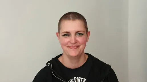 A photo of the author Lynda Clark standing in front of a white background. She is wearing black and has a shaved head.